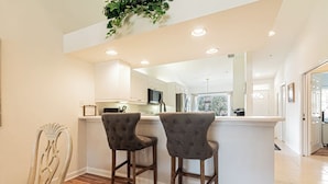 A modern kitchen with white cabinets, a central island, and two bar stools. - Well-appointed condo with a large lanai, outdoor kitchen and enclosed garage. (Naples)