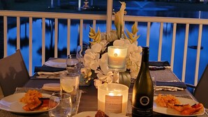 A dining table with plates of food, glasses, a candle, and a bottle of wine, set against a view of a body of water and palm trees. - Well-appointed condo with a large lanai, outdoor kitchen and enclosed garage. (Naples)