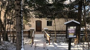 Lumipeitteinen polku johtaa rakennukseen, jossa on puuovi, ja sen lähellä on pieni rakennelma, jossa on kyltti.