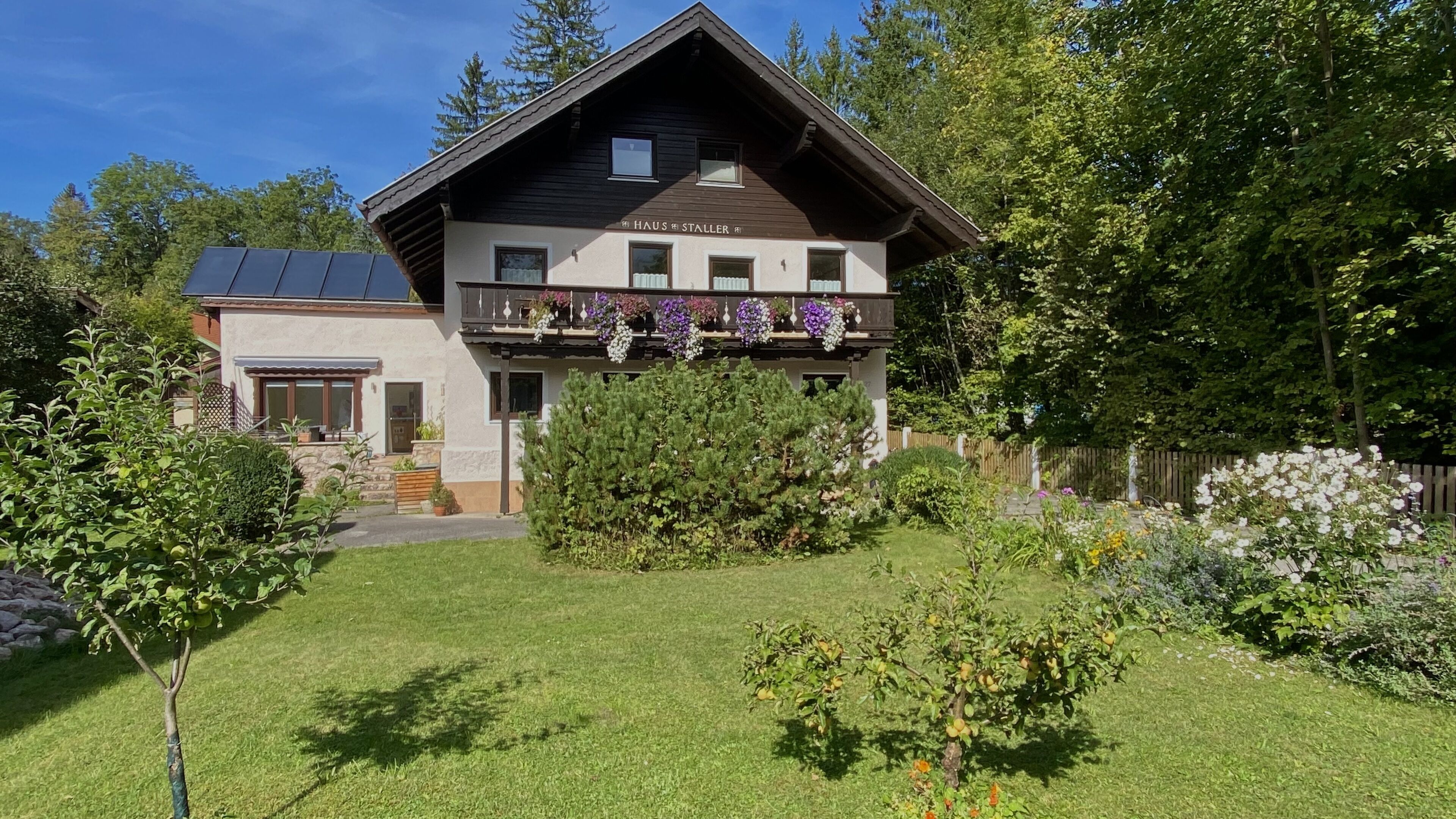 Un chalet à deux étages doté d’un balcon, entouré d’un jardin bien entretenu et d’une clôture en bois.