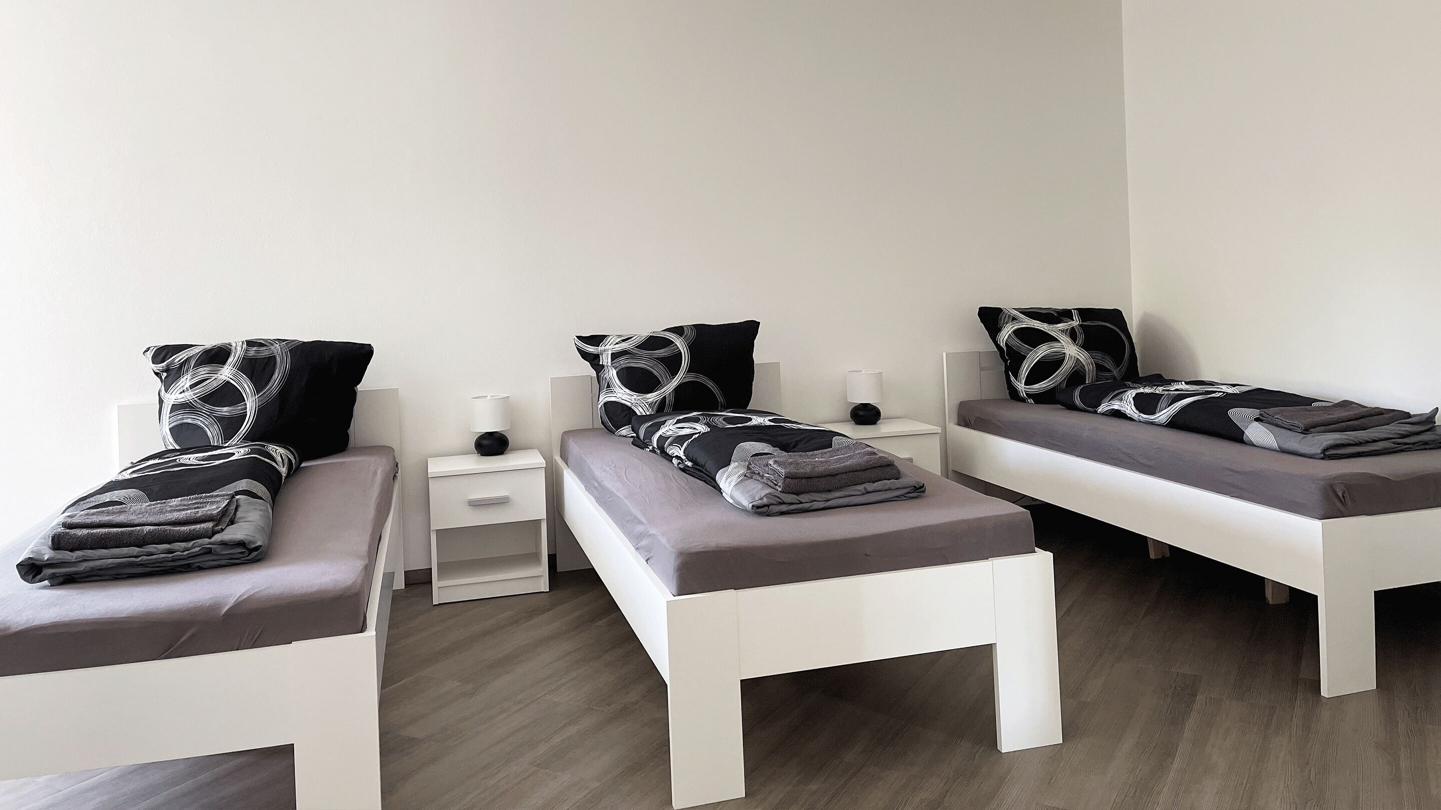 Une chambre d’hôtel avec trois lits simples, chacun étant équipé d’un oreiller et d’une couverture pliée. Il y a des tables de chevet avec des lampes et un petit tiroir.