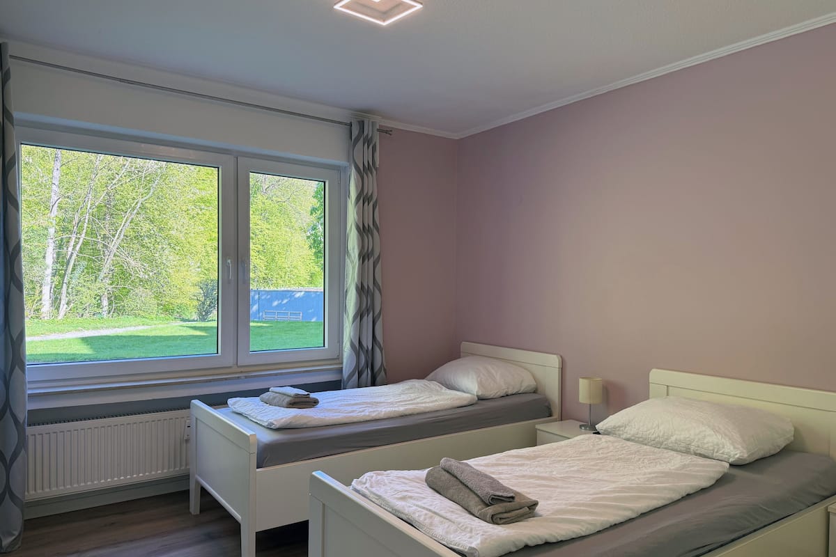 A bedroom with two beds, a window with a view of greenery, a radiator, and a bedside table with a lamp.