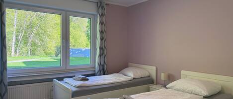 A bedroom with two beds, a window with a view of greenery, a radiator, and a bedside table with a lamp.