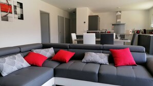 A modern living room with a grey sectional sofa, red and patterned cushions, a dining area with a white table and chairs, and a kitchen in the background.