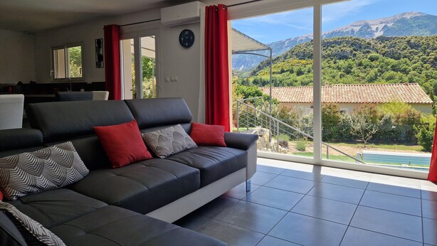 A modern living room with a grey sofa, red cushions, and a large window offering a mountain view.