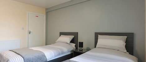 Habitación de hotel con dos camas individuales, una mesita de noche con lámpara y una puerta con cartel.