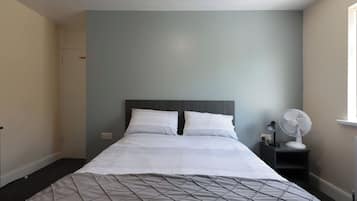 A neatly made bed with white linens and a quilted cover, a bedside table with a lamp and a fan, and a light blue wall.