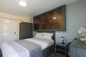 A bedroom with a bed, a wardrobe, a desk with a chair, a standing fan, and a large painting on the wall. - OYO Honeycroft Lodge, Uxbridge (Uxbridge)