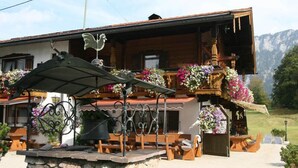 A chalet with a stone foundation, wooden balcony with flower baskets, outdoor seating, and a mountain backdrop. - Vacation apartment no. 14 for 2 - 4 persons, approx. 100 m², 1 bedroom, living room, kitchen, bathroom (Bischofswiesen)