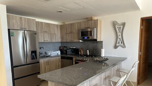 Una cocina moderna con un refrigerador de acero inoxidable, un microondas y una estufa. Hay armarios de madera y una encimera de granito. Una silla blanca está ubicada en la isla.