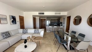 Una sala moderna con un sofá seccional, una mesa de comedor de vidrio con sillas, una mesa de centro de mármol y una cocina al fondo.