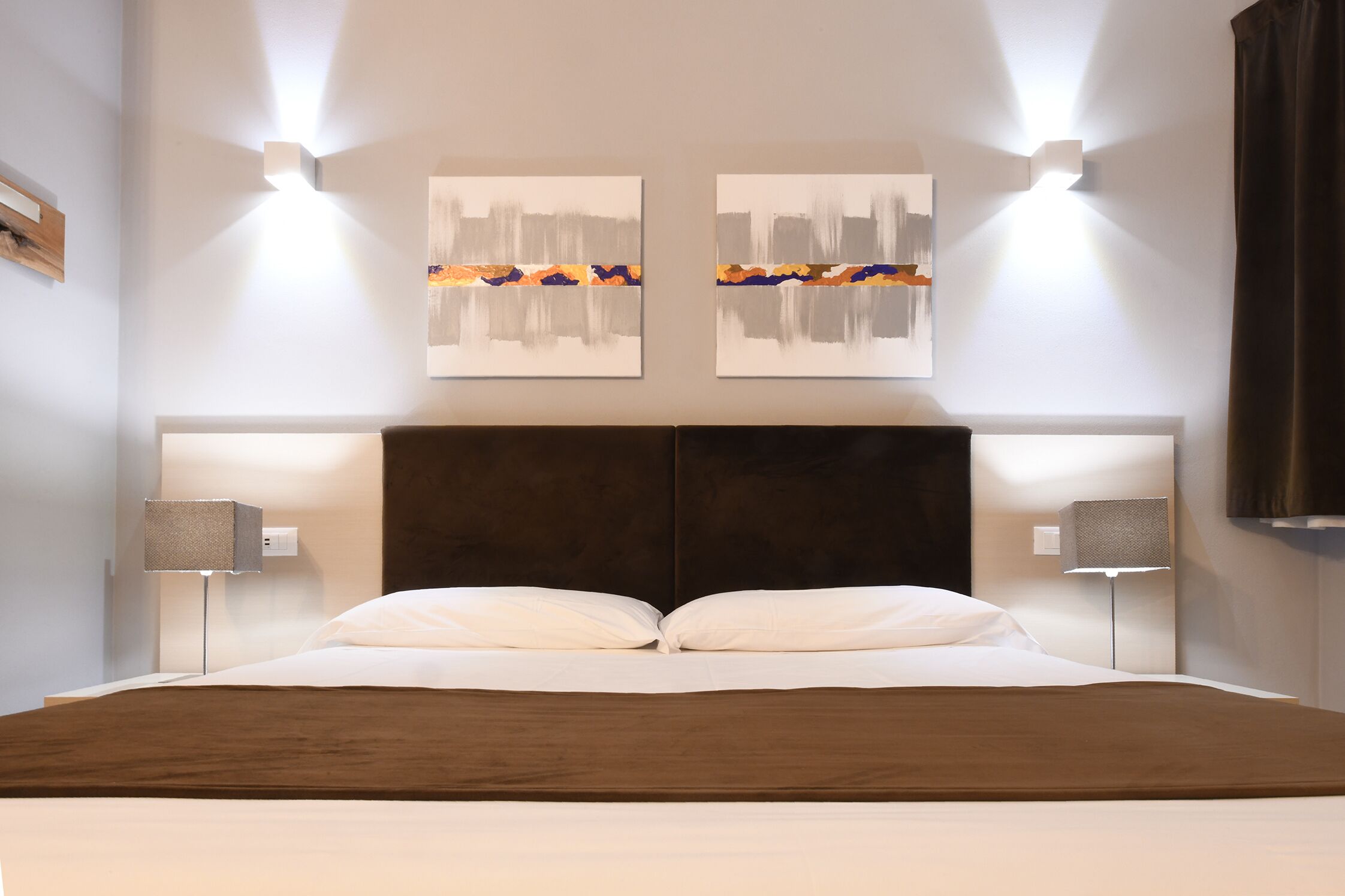 A modern hotel room with a large bed, two wall-mounted light fixtures, and abstract artwork above the bed.