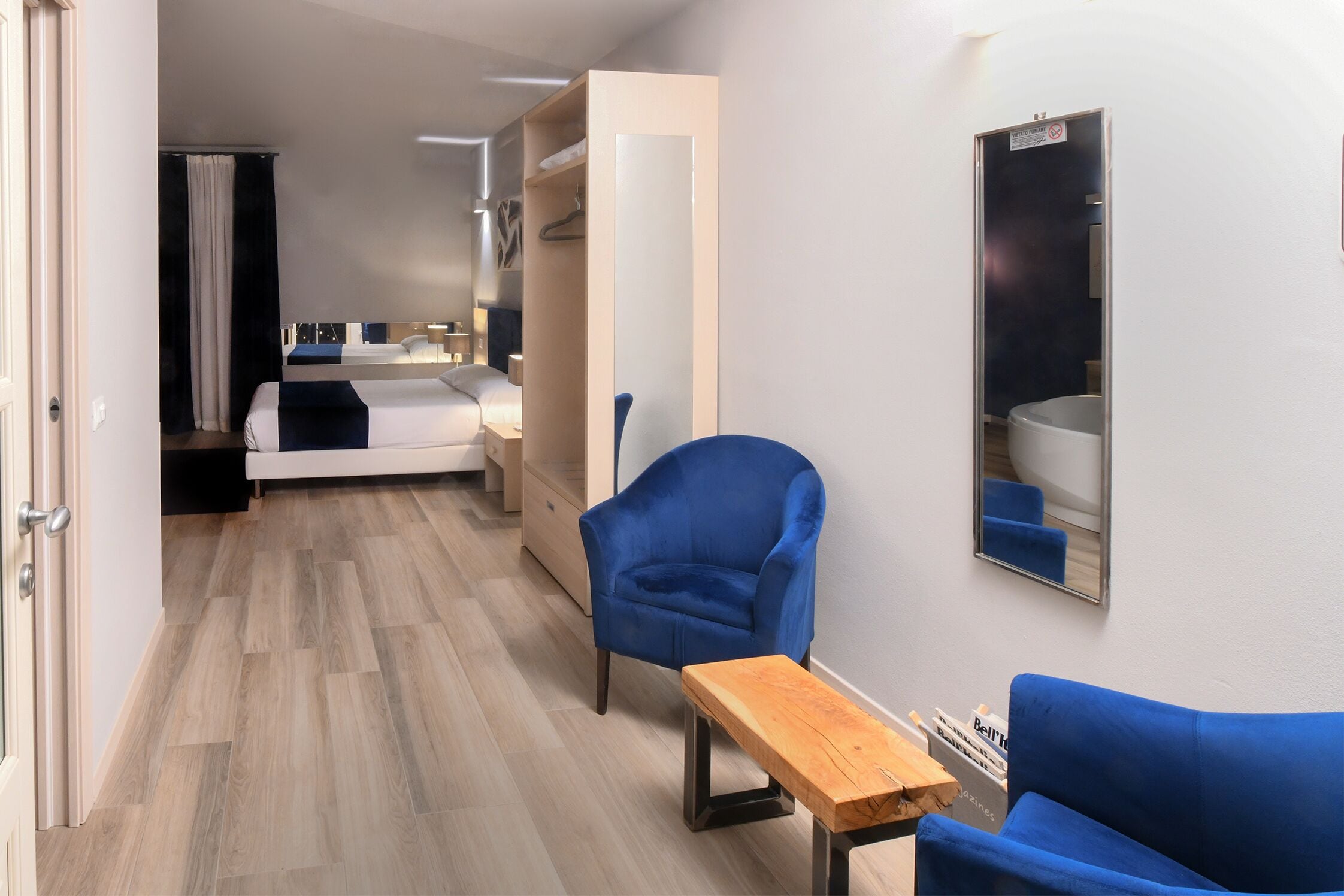 A modern hotel room with a bed, a blue armchair, a wooden table, and a mirror on the wall.