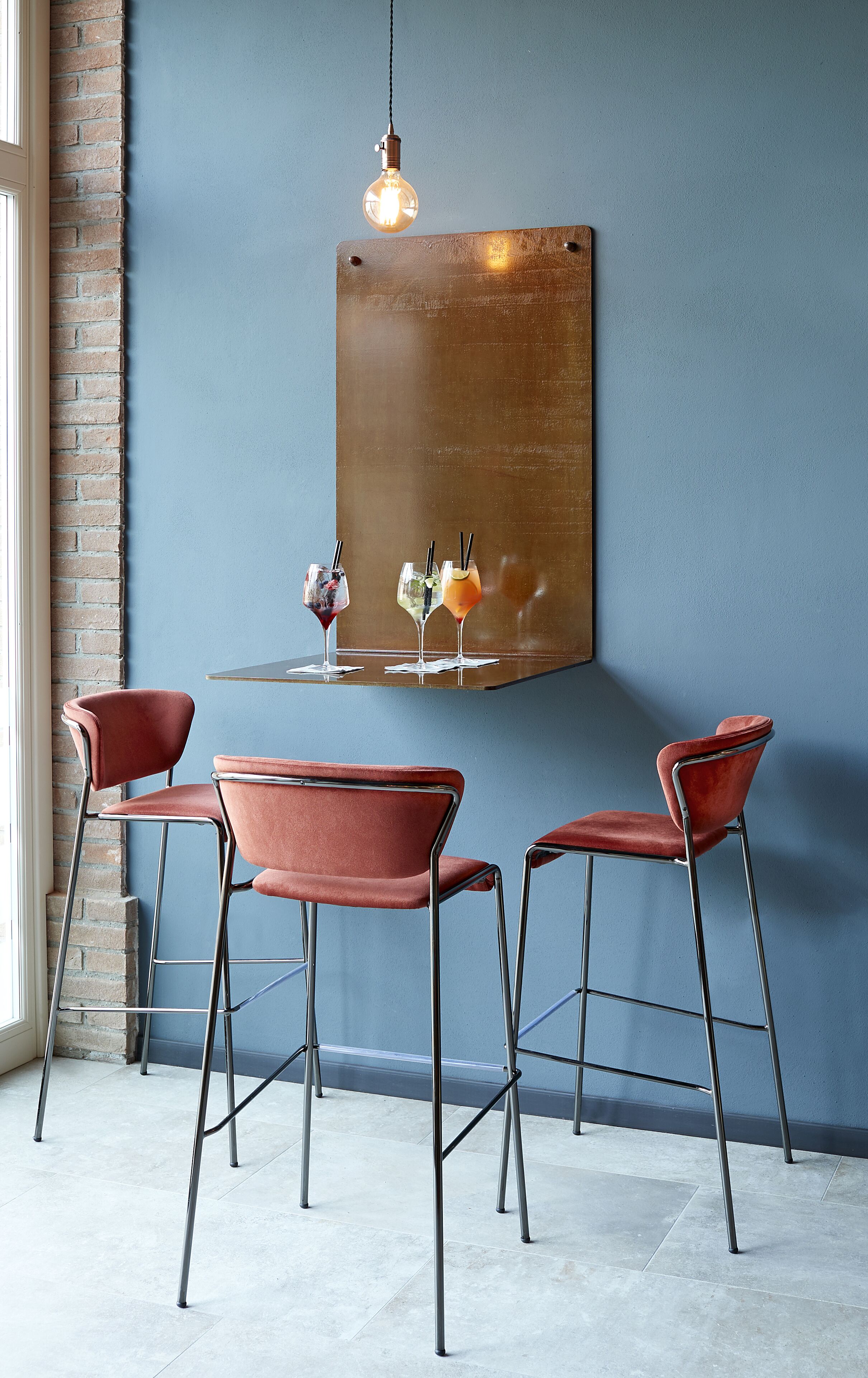 A dining area with a brick wall, a hanging light fixture, and three red chairs around a table with drinks.