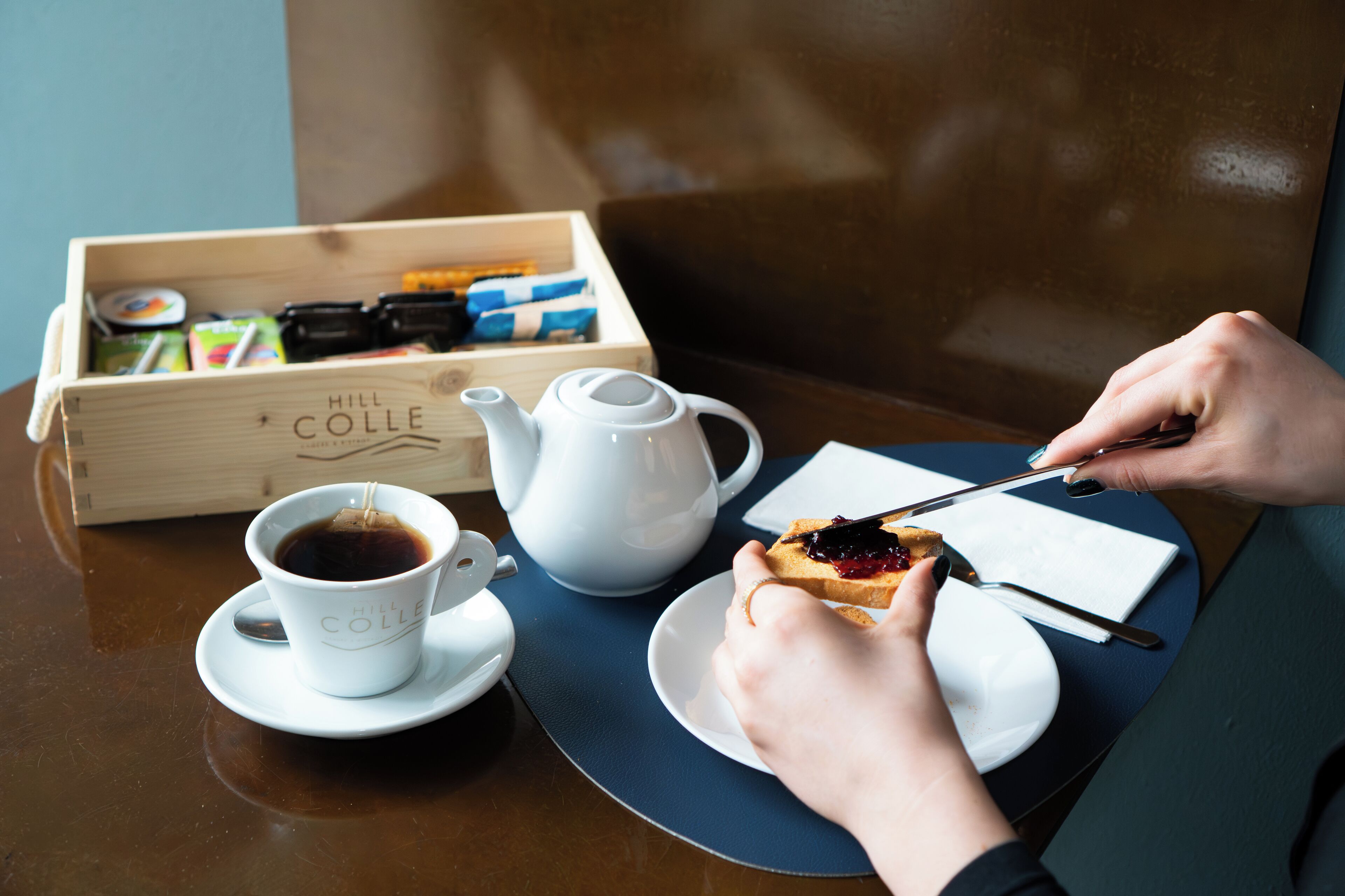 Een houten doosje met de tekst 'Hill Colle' waarin verschillende voorwerpen liggen, een witte theepot, een kop koffie, en een bord met toast en jam.