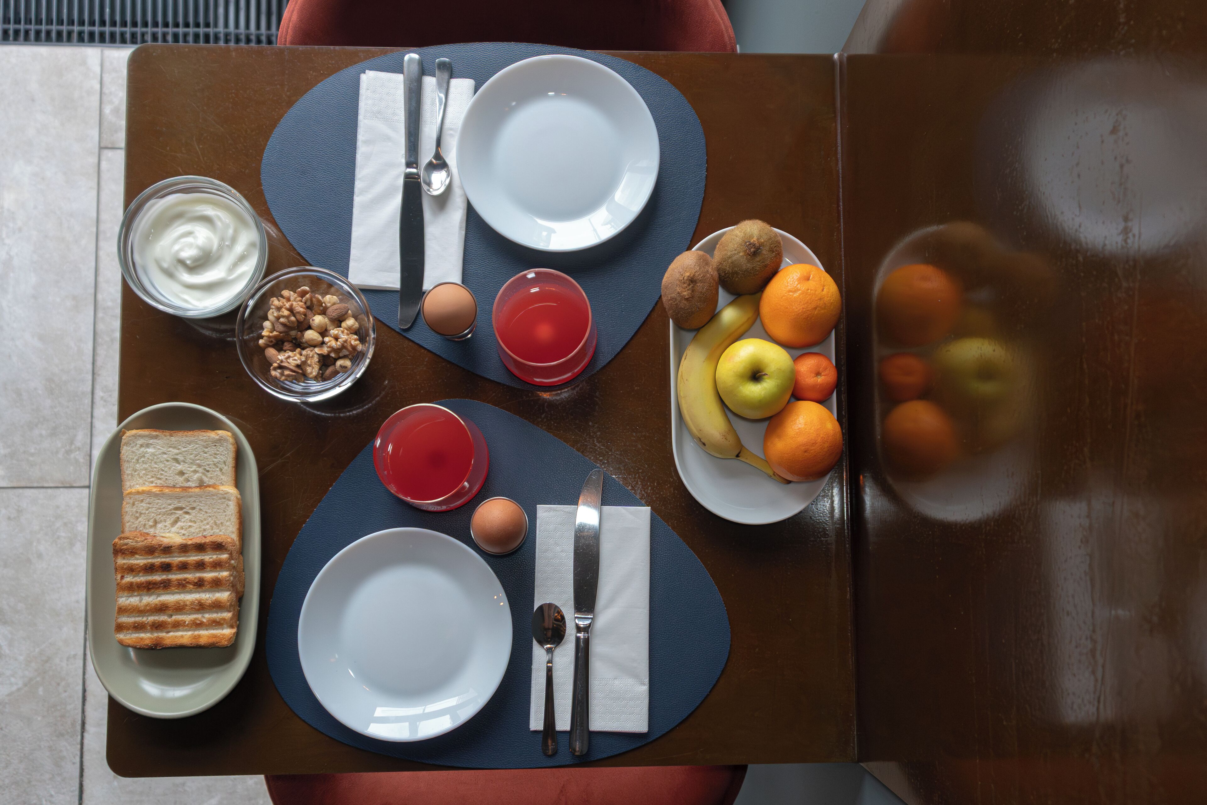 Une table dressée avec des assiettes, des couverts, un verre de jus de fruits, un bol de fruits, un pot de yaourt et une assiette de sandwichs au fromage grillé.