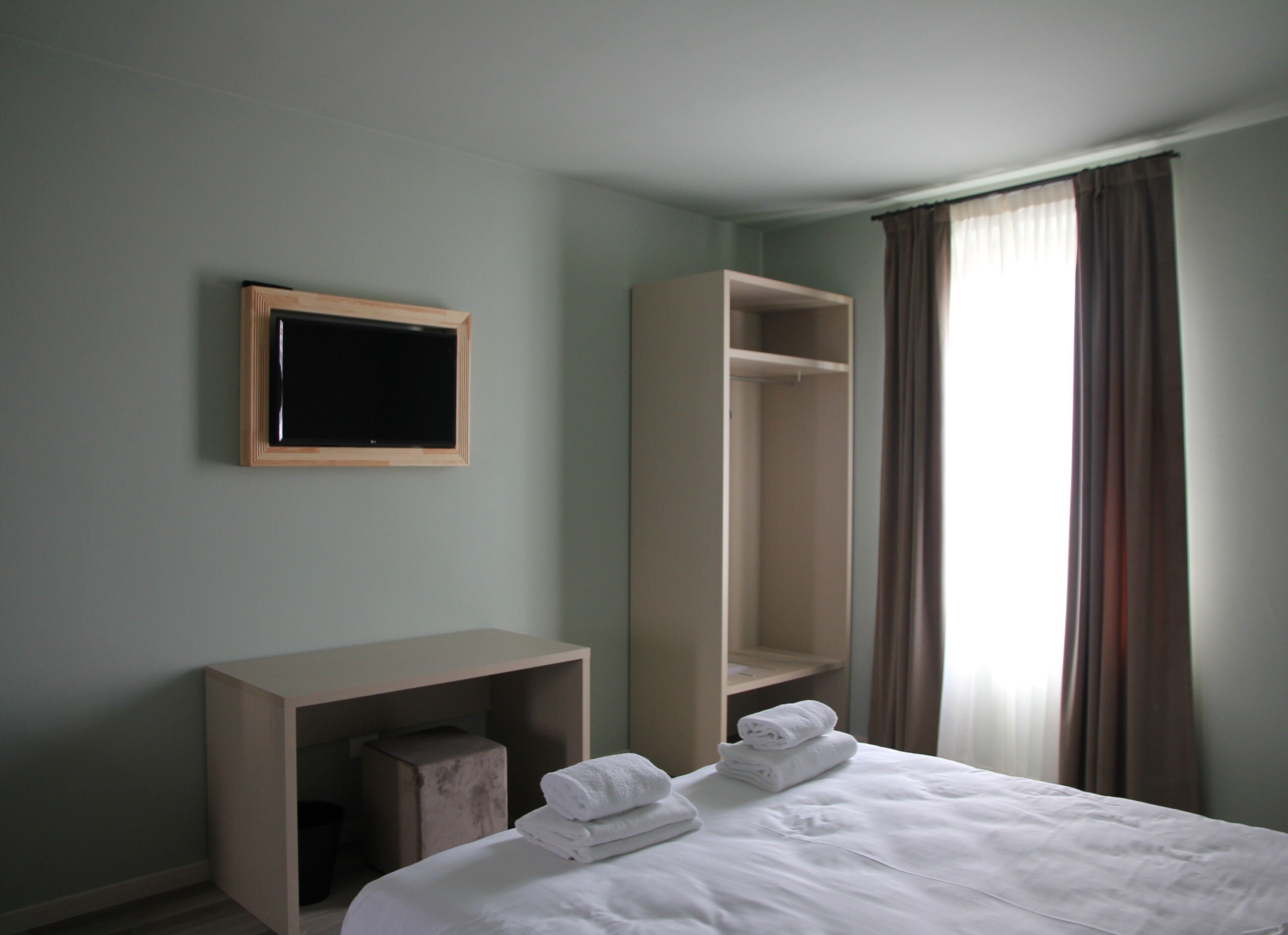 Una habitación de hotel con cama, mesita de noche, espejo, armario y ventana con cortinas.