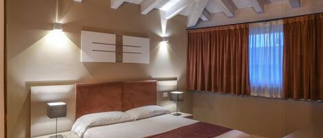 A bedroom with a bed, nightstands, wall-mounted lights, a window with curtains, and a sloped ceiling with exposed beams.