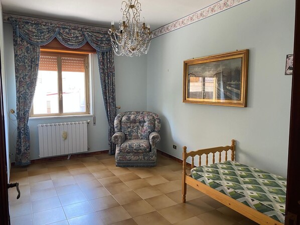 Ein Zimmer mit Sesseln mit Muster, einem Bett mit geometrischer Tagesdecke, einer Chandelüstere, einem gerahmten Bild an der Wand und einem Fenster mit Vorhängen.