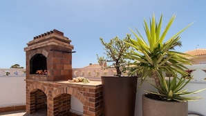 Ein gemauerter Außenofen mit Schornstein, eine Kübelpflanze mit Agave und eine geflieste Terrassenfläche.
