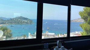 A room with a large window offering a view of a coastal town, a bed, a desk with a computer, and a window sill with bottled water and a kettle. - Life Butiq Hotel (Bodrum)