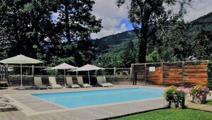 A rectangular swimming pool with lounge chairs and umbrellas, surrounded by a wooden deck and a grassy area. - Apartment Rose des Alpes 2 for 6 people 4 **** (Samoëns)
