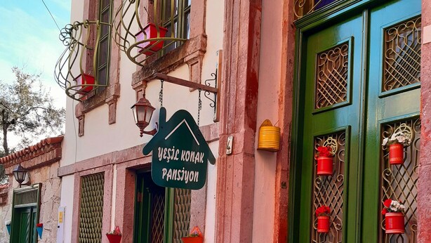 Un edificio con puerta verde, un letrero que dice "Yesil Konak Pansiyon" y macetas con flores rojas en los alféizares de las ventanas.