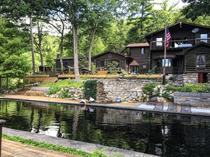 Un chalet rustique avec un mur en pierre et une terrasse en bois donnant sur un étang paisible.