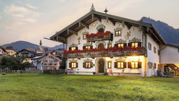 Ein traditionelles Gebäude im alpinen Stil mit einem Balkon, der mit roten Blumen geschmückt ist, einer Holztreppe und einem Blick auf die Berge.