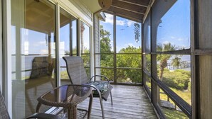 A covered outdoor area with a table and chairs, large windows, and a view of greenery and a bridge. - Whitman Point Townhome Unit A is Gulf Front with Sunsets & fishing pier (Cedar Key)