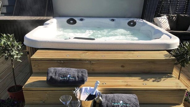 Un bain à remous avec des marches menant à celui-ci, entouré d’une terrasse en bois. Il y a deux coussins sur les marches et un seau contenant une bouteille et un verre.