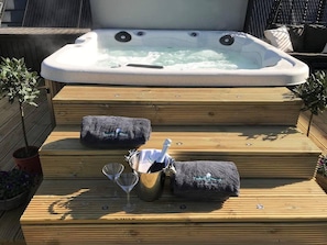 Un bain à remous avec des marches menant à celui-ci, entouré d’une terrasse en bois. Il y a deux coussins sur les marches et un seau contenant une bouteille et un verre.