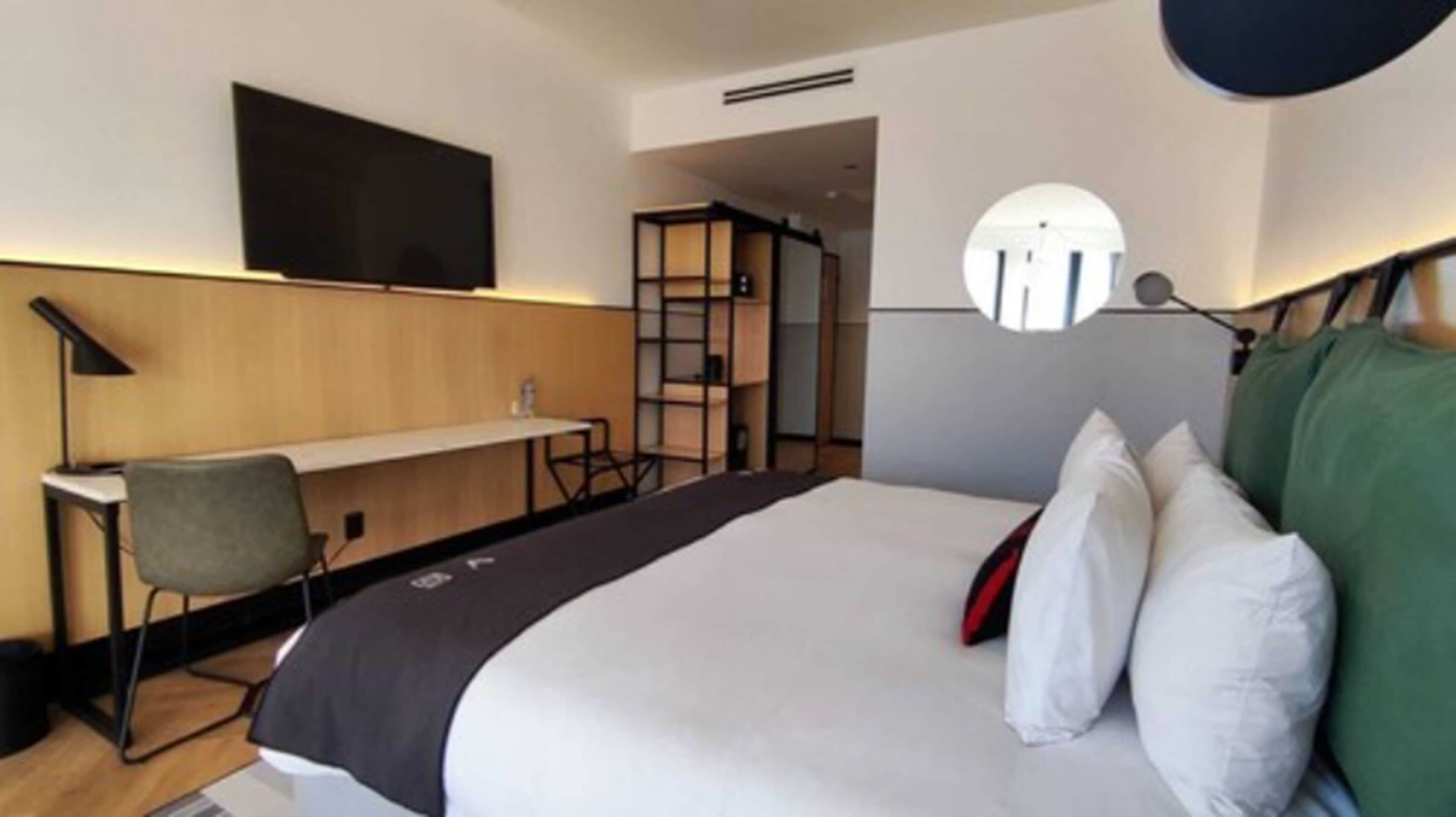 Quarto de hotel com cama, escrivaninha, cadeira, televisão e espelho redondo.