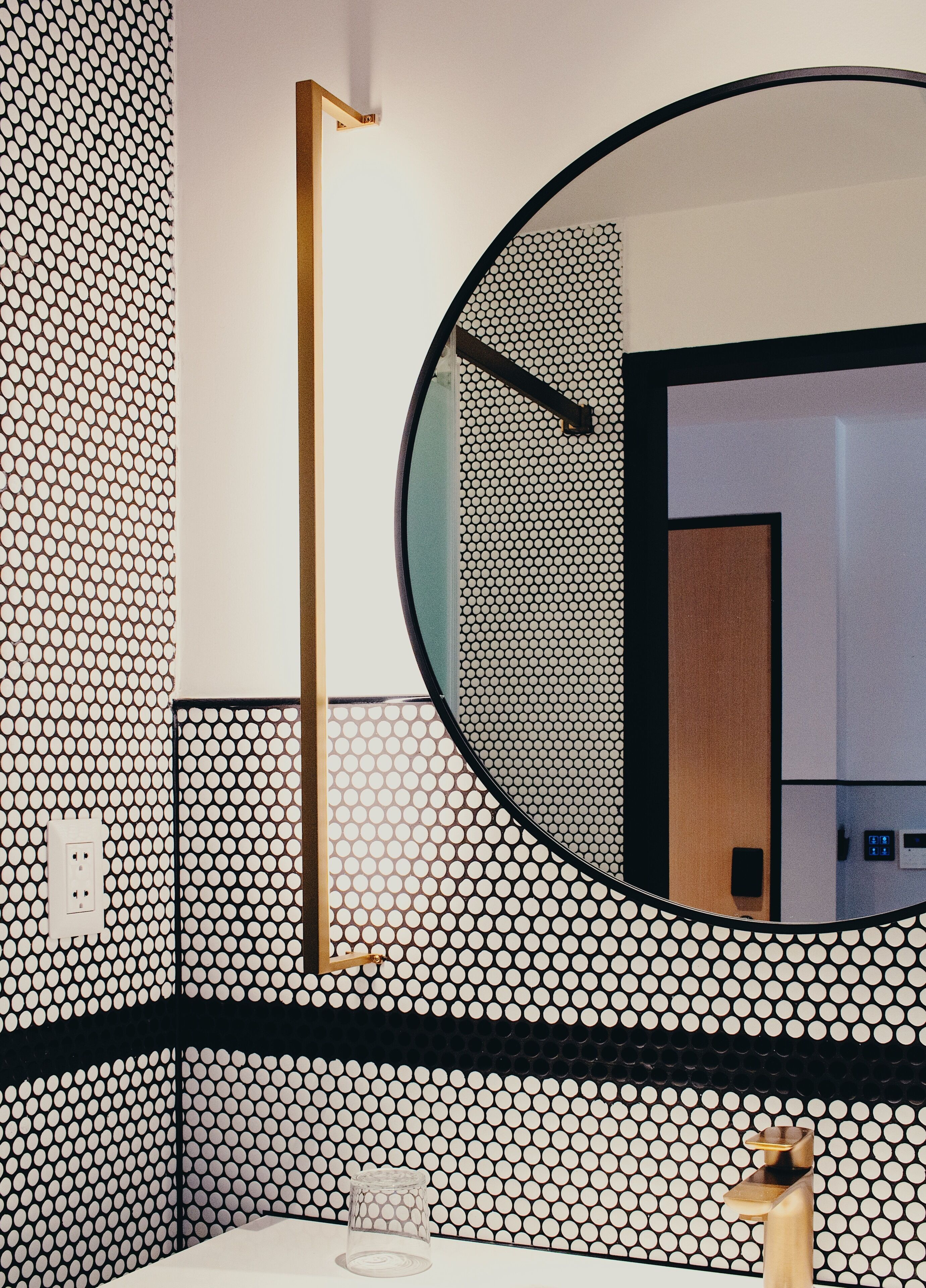 Un baño con espejo redondo, pared con estampado blanco y negro, y grifo dorado.