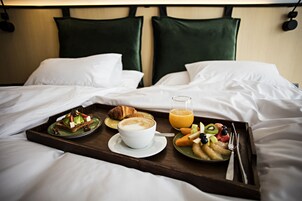 Una bandeja de desayuno ordenada sobre una cama con sábanas blancas, que incluye croissants, fruta, un vaso de jugo de naranja y una taza de café.