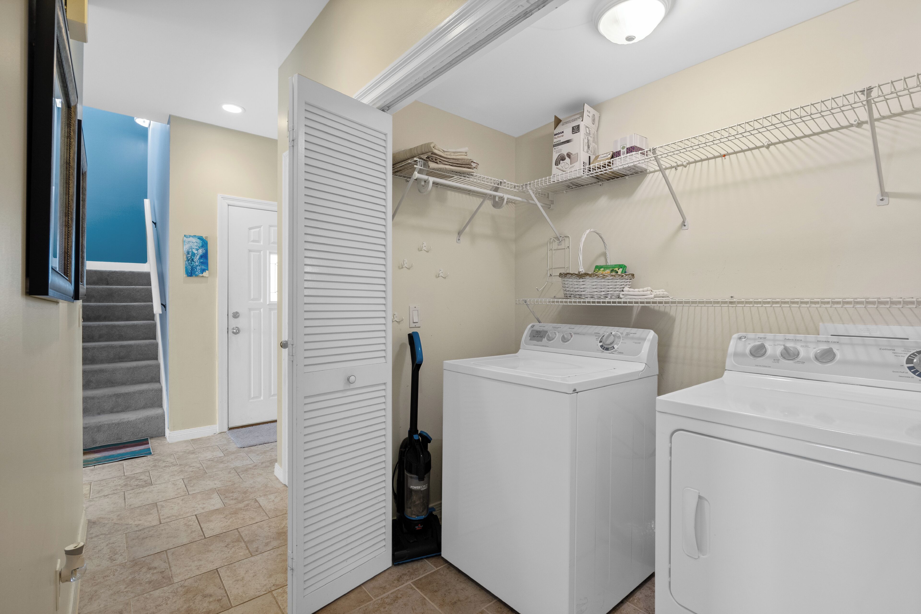 A laundry room with two washing machines, a dryer, a ceiling-mounted shelf with folded towels, and a vacuum cleaner.
