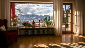Ein gemütliches Zimmer mit großem Fenster und Blick auf die Berge, einem roten Sessel, einer Heizung und einer Topfpflanze.