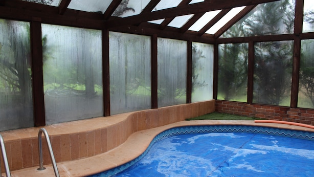 An indoor swimming pool with a tiled edge, a metal ladder, and a glass enclosure with a view of trees. - Cabañas Esperanza (Tlalpujahua)