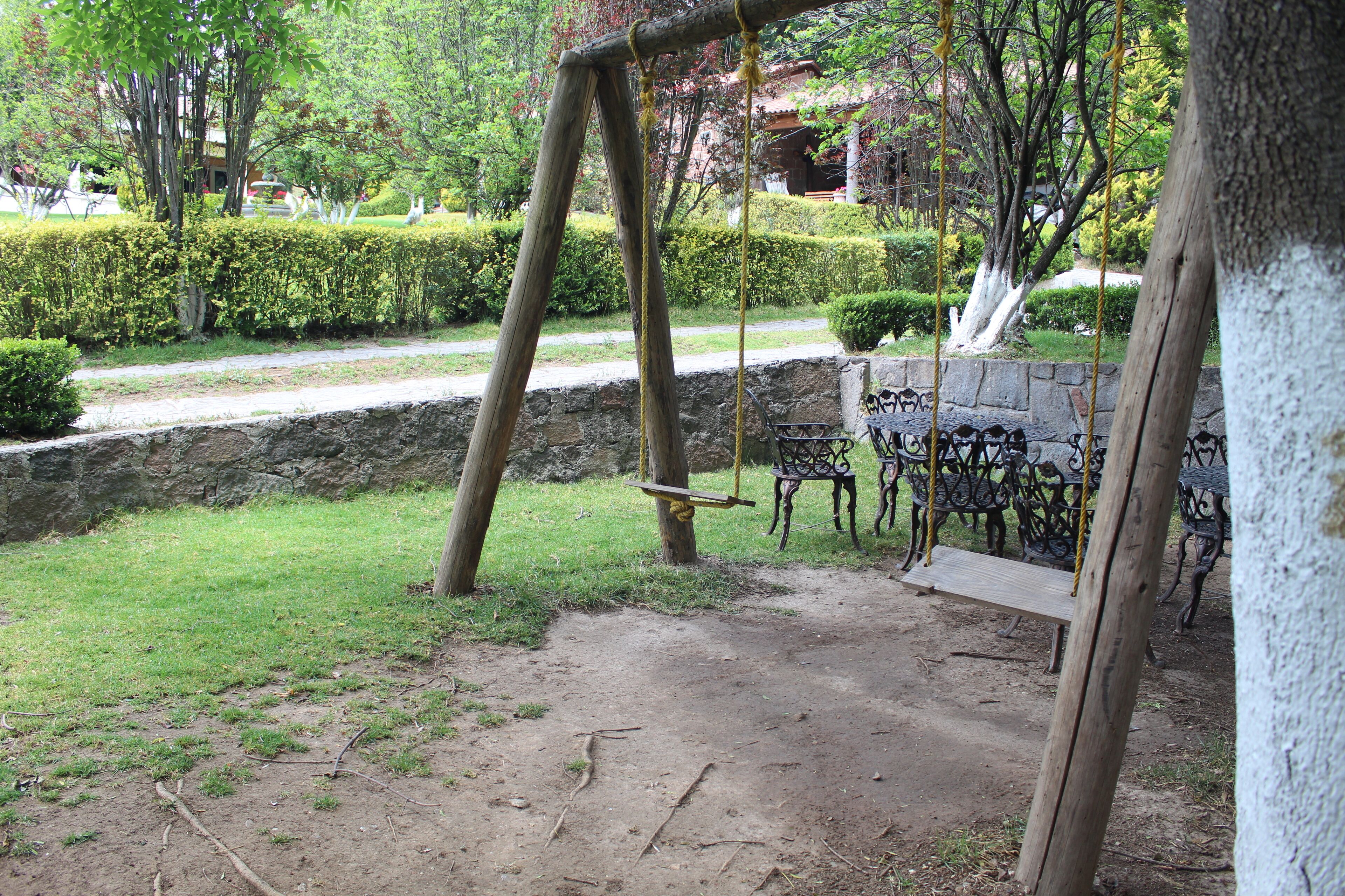 Un conjunto de columpios de madera en un jardín con un muro de piedra y bancos de metal.