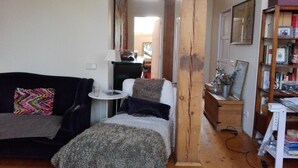 Een gezellige woonkamer met een bank, een klein tafeltje met een lamp, een boekenkast en houten balken.