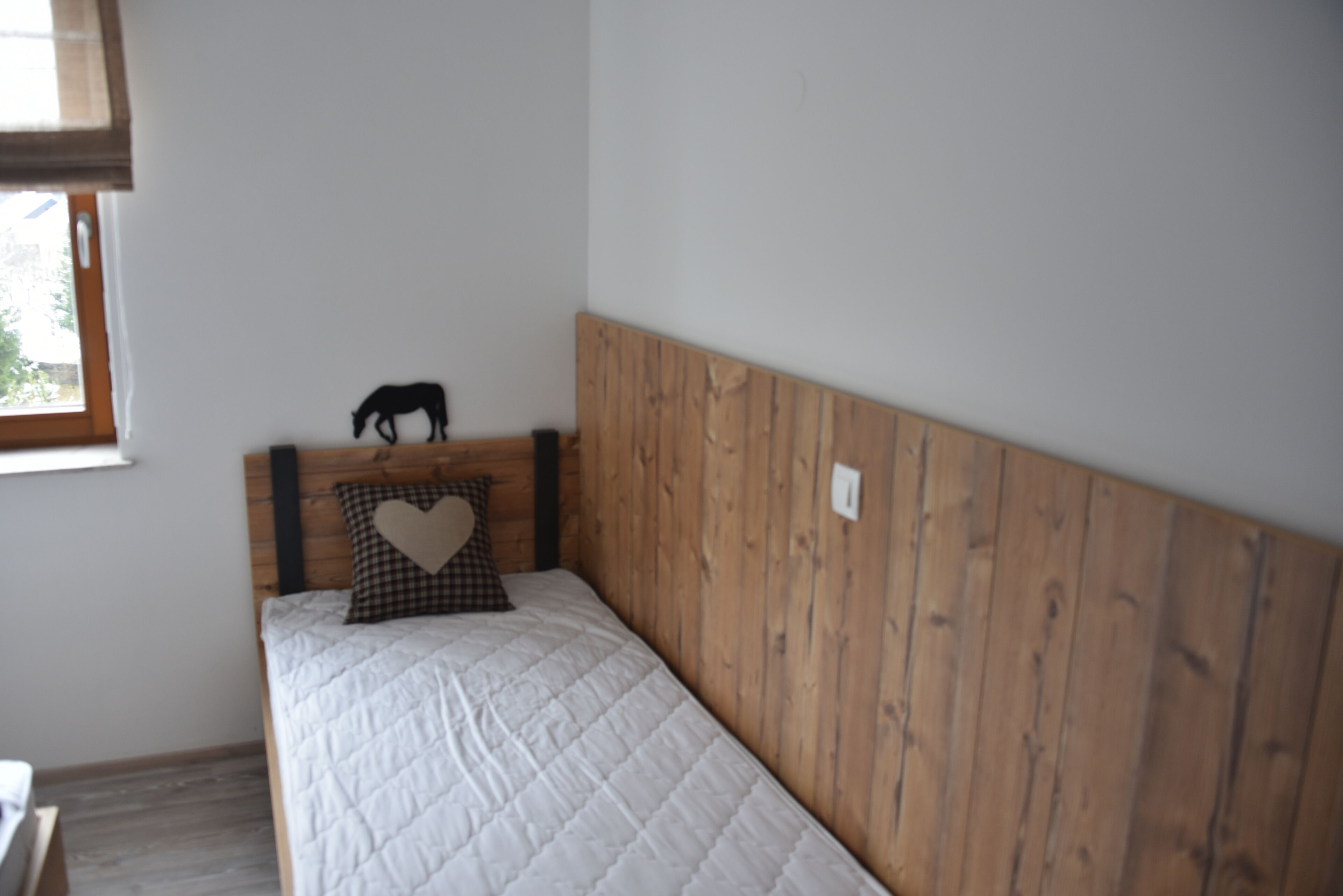 Een enkel bed met een houten hoofdbord, een hartvormig kussen en een klein zwart silhouetdecoratie op het hoofdbord.