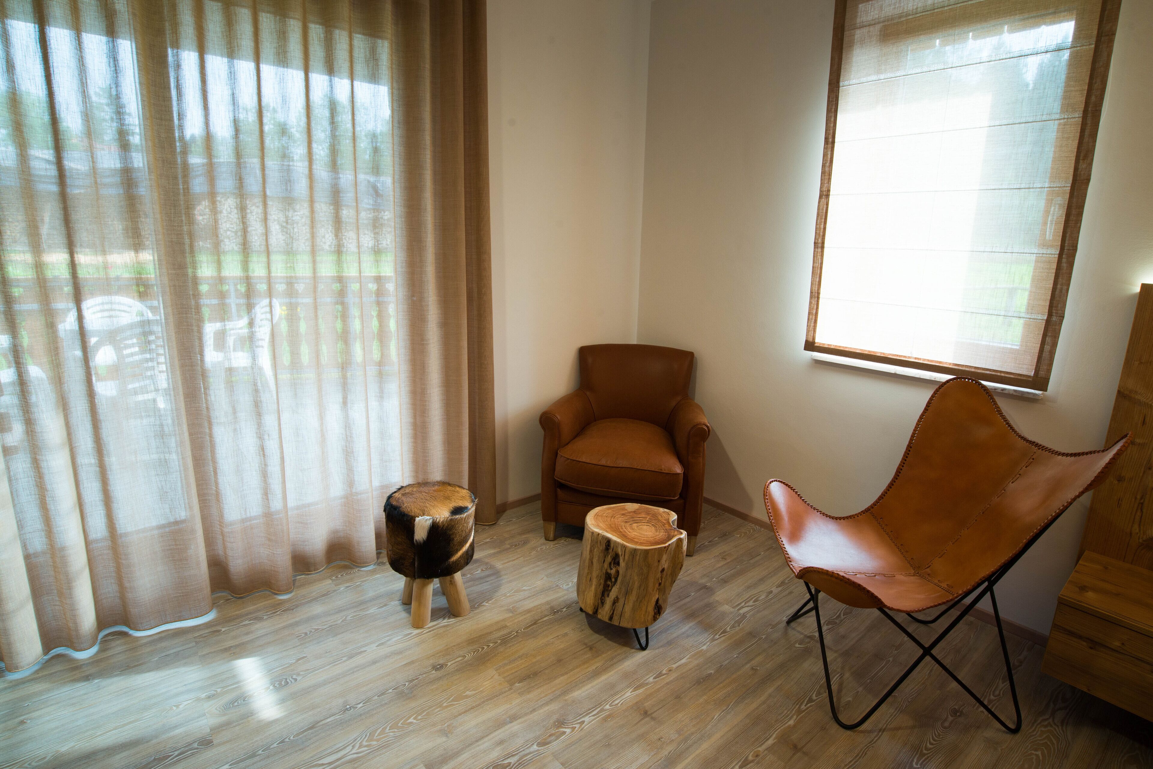 A room with a wooden floor, a brown leather chair, a small wooden stool, and a window with sheer curtains.