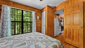 Un dormitorio con un ventanal grande, paredes de madera, una cama con colcha y vistas a una cocina.