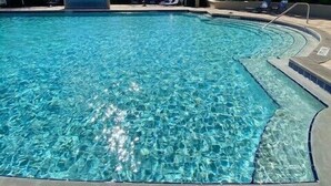 Une piscine d’eau cristalline, d’un bleu éclatant, sous un ciel ensoleillé, entourée de palmiers et de bâtiments.