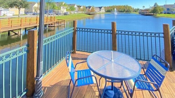 Une terrasse en bois avec une table ronde en verre et des chaises bleues, donnant sur un lac paisible sous un ciel bleu clair.