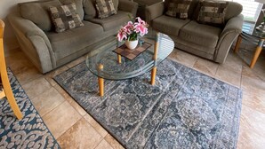 Un salon comprenant un ensemble de canapés, une table basse en verre agrémentée d’un arrangement floral, une lampe et une grande fenêtre avec des stores.