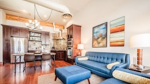 Una sala moderna con un sofá azul, un comedor con mesa y sillas, y una cocina con armarios oscuros y electrodomésticos de acero inoxidable.