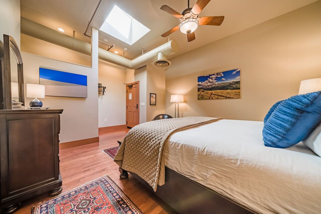 Une chambre à coucher avec un lit, un ventilateur de plafond, un puits de lumière, une commode en bois, un tapis et un tableau accroché au mur.