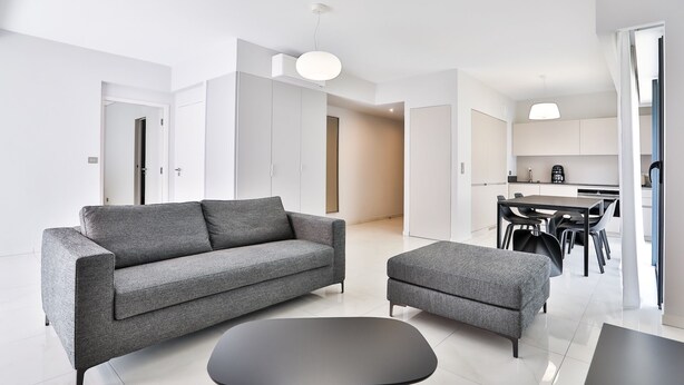Een moderne woonkamer met een grijze bank, een bijpassend voetenbankje en een zwarte salontafel. In de achtergrond is een eethoek met een zwarte tafel en stoelen zichtbaar.