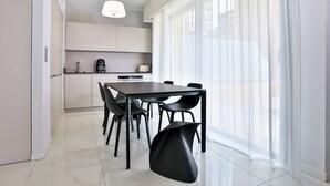 Een moderne keuken met een eethoek, met een groot raam voorzien van dunne gordijnen, een hanglamp en een strakke zwarte eettafel met stoelen.